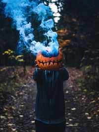 Man holding smoke emitting pumpkin while standing in forest