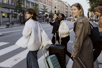 Young female friends crossing road while pulling luggage in city