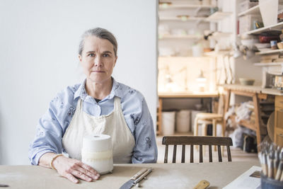 Portrait of confident senior female potter sitting at table with vase against white wall in workshop