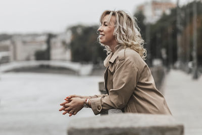 A middle-aged blonde woman with curly hair stands on the river embankment in the city.