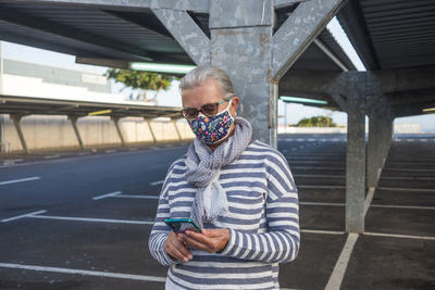 Senior woman using smart phone wearing mask standing outdoors