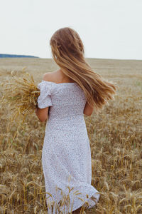 Woman in rye field wearing white dress with wind in hair, back view