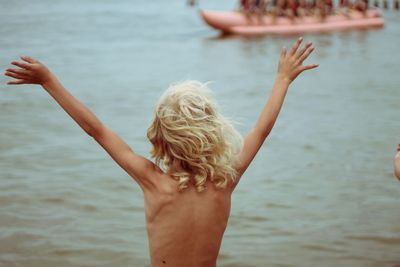 Rear view of shirtless boy waving hands while standing by water