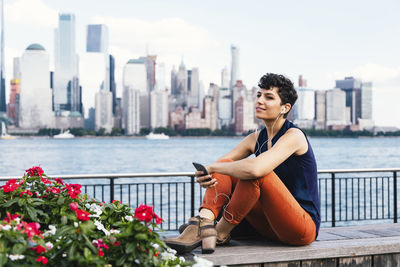 Woman listening music while sitting on retaining wall by river against buildings in city