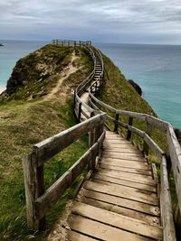 Wooden railing on footpath by sea against sky