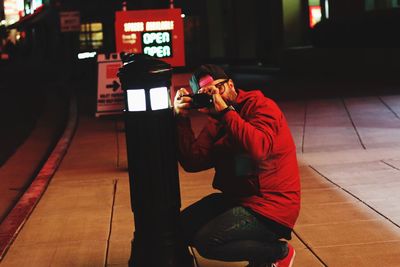 Man photographing illuminated bollard with mobile phone sitting on sidewalk at night