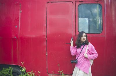 Young woman holding mobile phone while standing against red wall