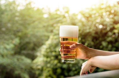 Cropped hand of man holding beer glass against trees