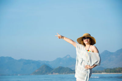Portrait of young woman gesturing peace sign while standing at beach against clear sky