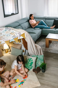 Relaxed mother using television remote control sitting on sofa while children playing with game