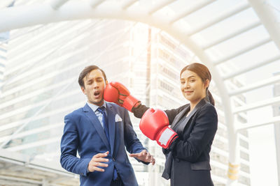 Portrait of smiling young businesswoman hitting colleague with red boxing gloves against buildings in city