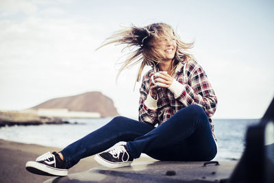 Cheerful young woman tossing hair on vehicle at beach against sky