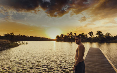 Portrait of shirtless man standing on jetty over lake against sky during sunset
