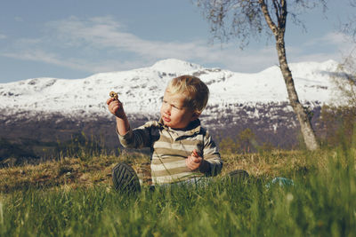 Cute boy sitting on grassy land against snowcapped mountain