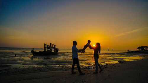 Parents holding baby while standing at beach against sky during sunset