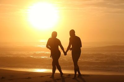 Silhouette couple holding hands and walking at beach against sky during sunset