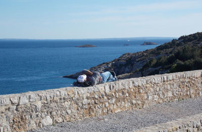 Man resting on retaining wall by sea against sky