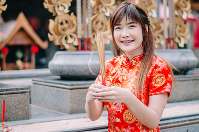 Portrait of smiling woman in traditional clothing holding incense sticks standing against built structure
