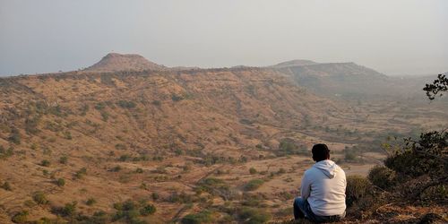Rear view of young man sitting on mountain against sky