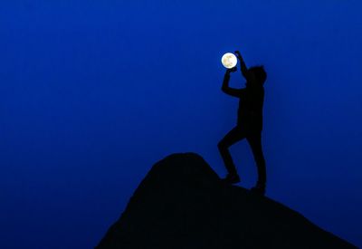 Optical illusion of silhouette man holding moon against blue sky at dusk