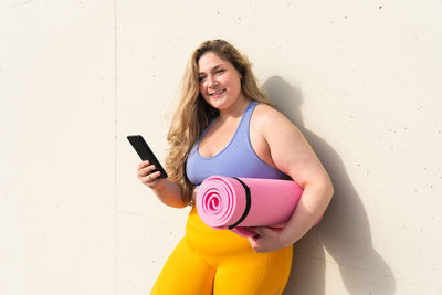 Portrait of young woman with mobile phone standing against wall