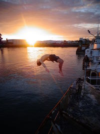 Full length of young man jumping in sea against cloudy sky during sunset