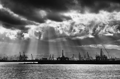 Cranes at harbor by sea against cloudy sky