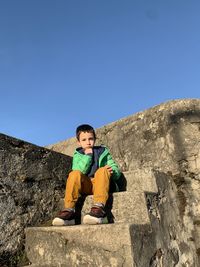 Full length of boy sitting on rock against clear blue sky