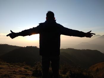 Rear view of man with arms outstretched standing against sky during sunset