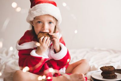 Cute child girl 3-4 year old wear red santa claus hat and bathrobe sit in bed with biscuit 