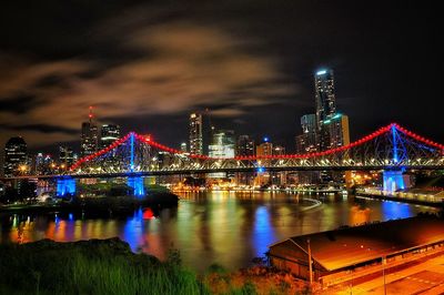 Illuminated bridge over river against buildings at night