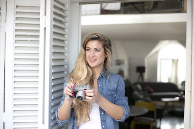 Smiling young woman holding camera by door at home