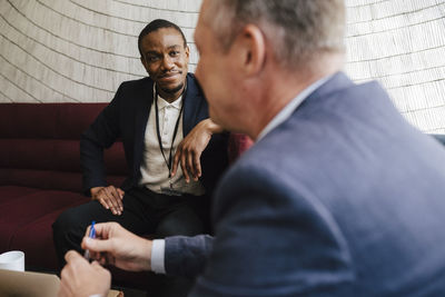 Smiling businessman looking at male colleague discussing while sitting on sofa during networking event