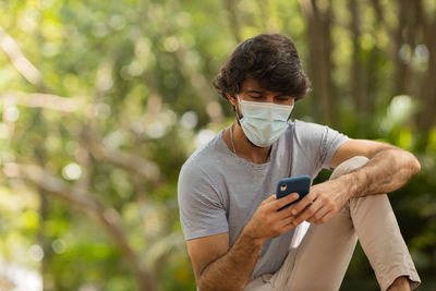 Young man wearing a mask and a smartphone at day time at a green park . mobile phone, technology