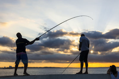 Rear view of man fishing in sea against sunset sky