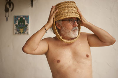Portrait of shirtless man wearing hat standing against wall