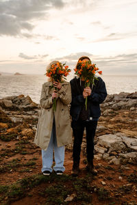 Full body anonymous boyfriend and girlfriend in outerwear covering faces with fresh bouquets while standing on seashore at sunset in aviles, spain