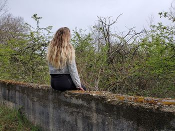 Rear view of young woman sitting on retaining wall against plants