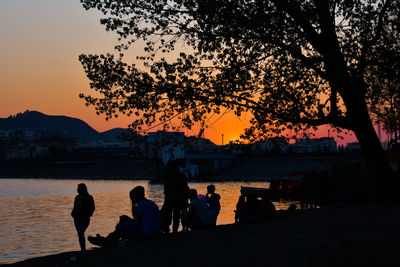 Silhouette people sitting on shore against sky during sunset