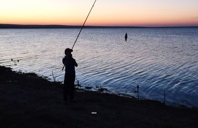 Silhouette man fishing in sea against sunset sky