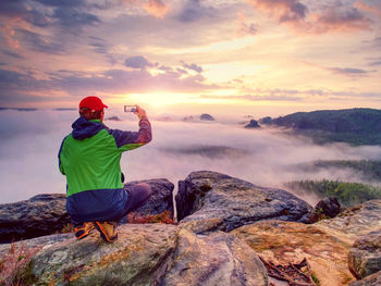 Hiker sit on heels and takes selfie photo, fall nature adventure. man sit on stone on mountain sumit