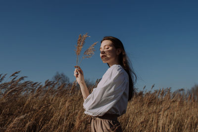Young woman in white blouse standing in field and holding dry pampas grass in front of sky