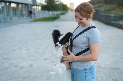 Happy caucasian woman walking with a dog in a backpack. papillon spaniel continental in a sling