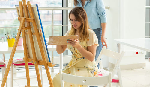Young woman painting while woman standing in background