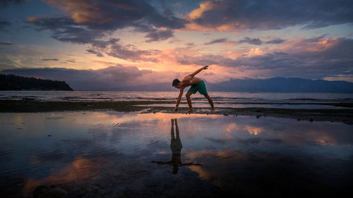 Shirtless man on shore at beach against sky during sunset