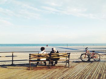 Rear view of women sitting on park beach at beach against cloudy sky