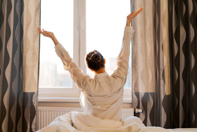 Back view caucasian woman stretching arms and body near window after waking up