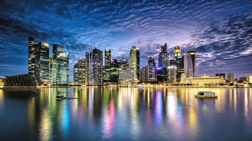 Illuminated skyscrapers in city by river against sky at dusk