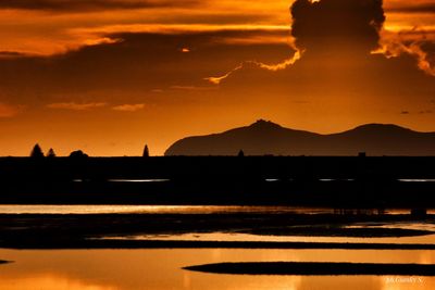 Scenic view of silhouette mountains by lake against orange sky