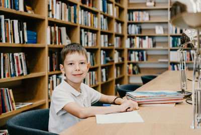 The boy sits in the library and writes in a notebook at the table. preparing for homework. 